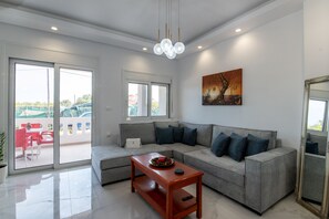 A modern living room with a grey sectional sofa, a wooden coffee table, and a large window with a view of a balcony. - Stella s House Near the Sea in Tavronitis (Platanias)