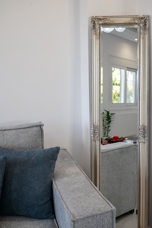 Ein Zimmer mit grauem Sofa, einem großen, verzierten Spiegel und Blick auf ein Fenster und eine Heizung.