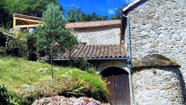 A stone building with a tiled roof, a wooden structure in the background, and a garden with hydrangea flowers. - Mas du Roy (Sénéchas)