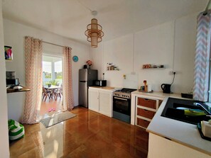 Eine moderne Küche mit Kühlschrank, Herd, Mikrowelle und Backofen. Ein Essbereich mit Stühlen und Tisch bietet einen Blick ins Freie durch eine Tür.