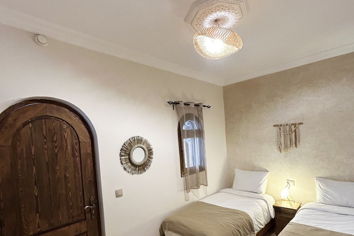 A room with two beds, a wooden door, a mirror, a ceiling light fixture, and a patterned floor.