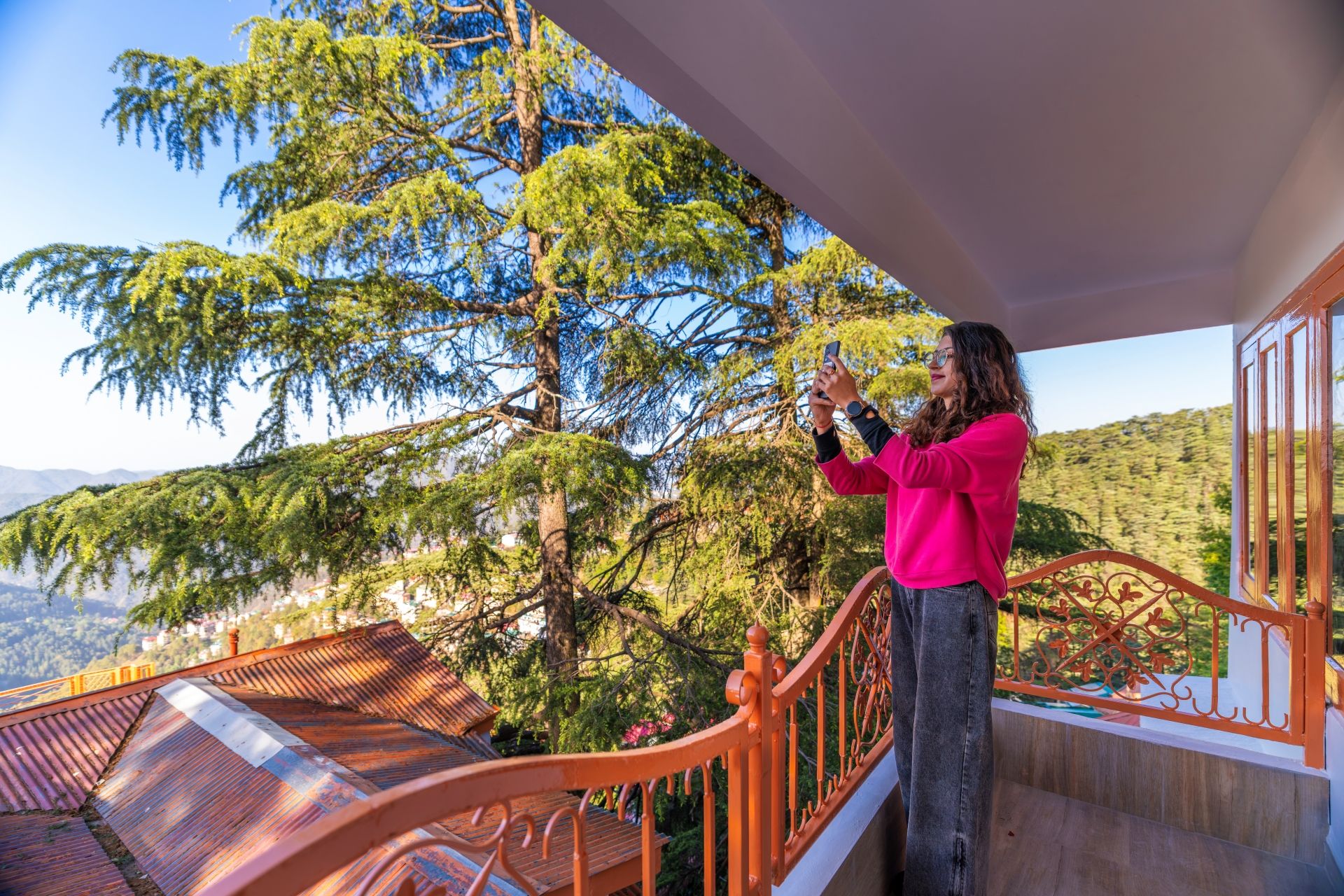Una persona tomando una foto en un balcón con vista a un árbol grande y un paisaje montañoso.