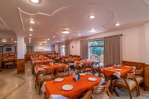 A spacious dining area with multiple tables and chairs, a high ceiling with recessed lighting, and a view of the outdoors through large windows.