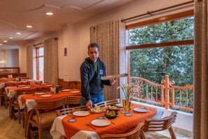 A dining area with tables set for meals, a waiter holding a tray, and a view of trees through large windows. - Kalra Regency Shimla (Shimla)