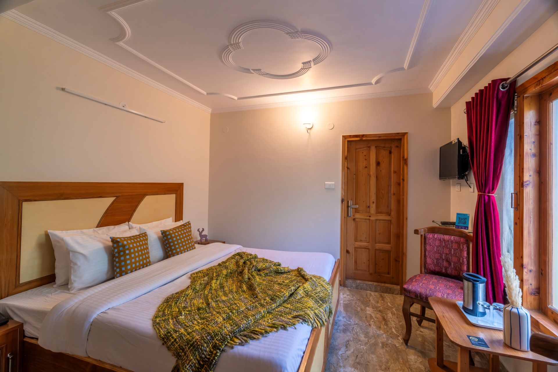 A bedroom with a bed, a wooden headboard, a bedside table, a chair, a window with curtains, and a television on the wall.