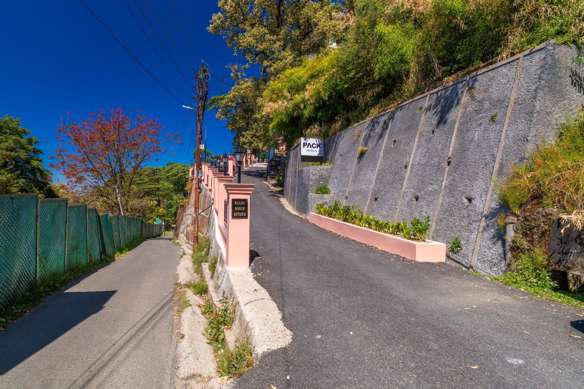 Una carretera estrecha con una pendiente pronunciada, bordeada por una cerca verde y un muro de piedra. Se observan letreros que indican 'PACK SHIMLA' y 'KALRA HOUSE ESTATE'.