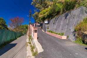 Una carretera estrecha con una pendiente pronunciada, bordeada por una cerca verde y un muro de piedra. Se observan letreros que indican 'PACK SHIMLA' y 'KALRA HOUSE ESTATE'.