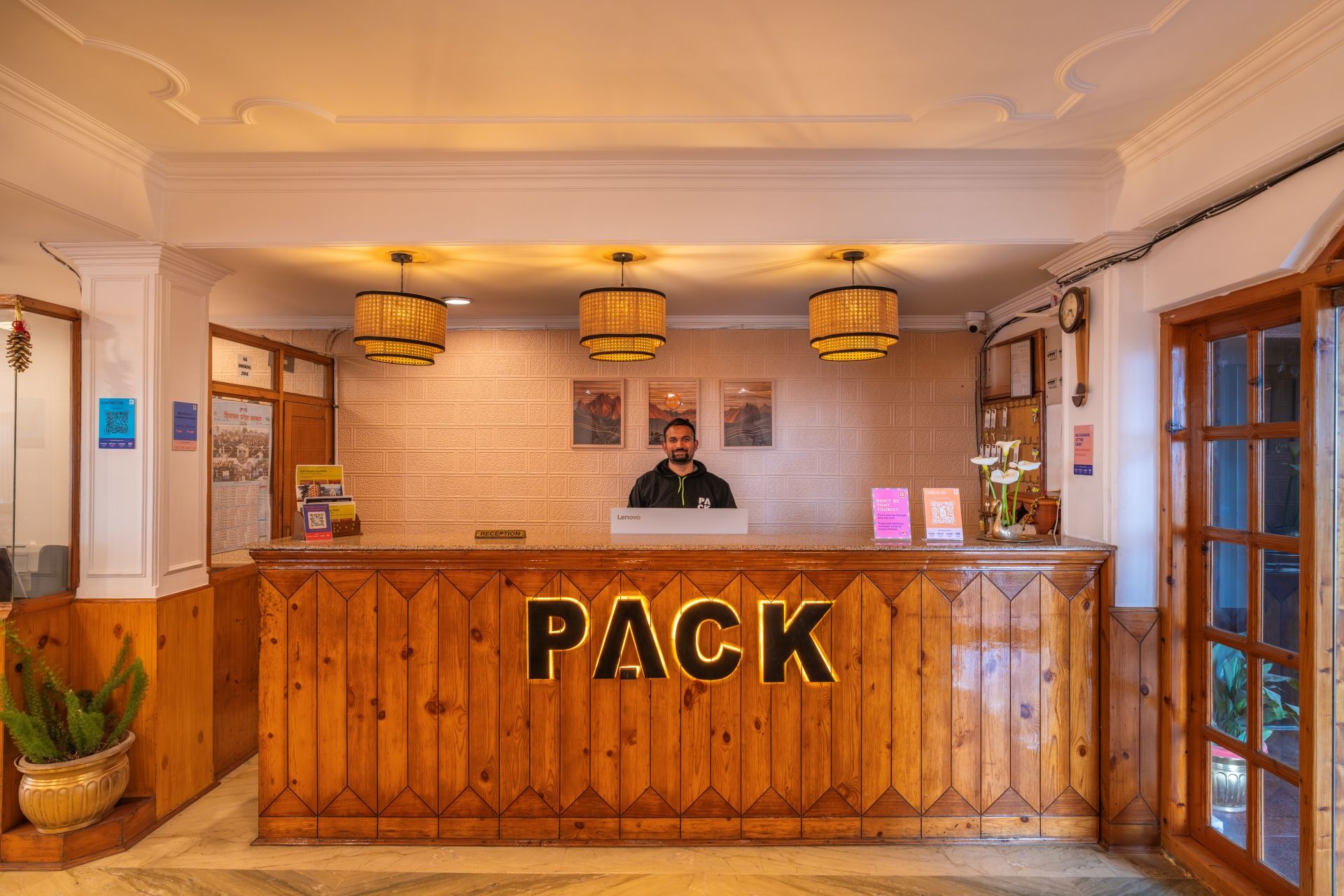 Mostrador de recepción con el rótulo 'PACK', acabado de madera, tres lámparas colgantes y una recepcionista.