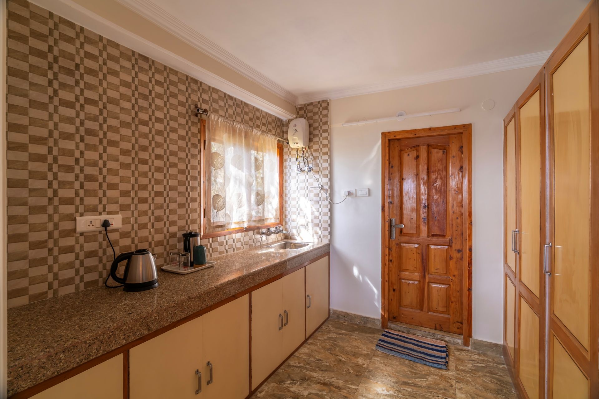 Una cocina con salpicadero a cuadros, una ventana con cortina de hojas, una puerta de madera y piso de baldosas.