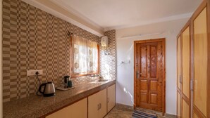 A kitchen with a checkered backsplash, a window with a leaf-patterned curtain, a wooden door, and a tiled floor. - Kalra Regency Shimla (Shimla)