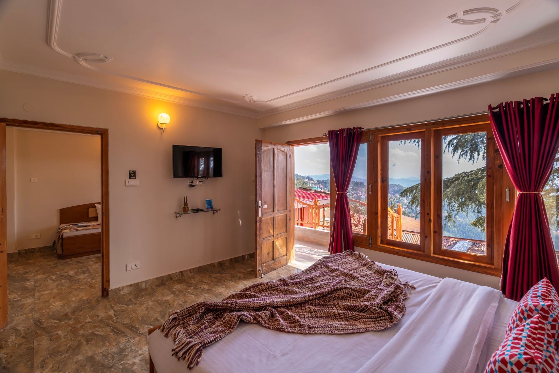 A bedroom with a bed, a TV, a shelf with items, and a view through a window.