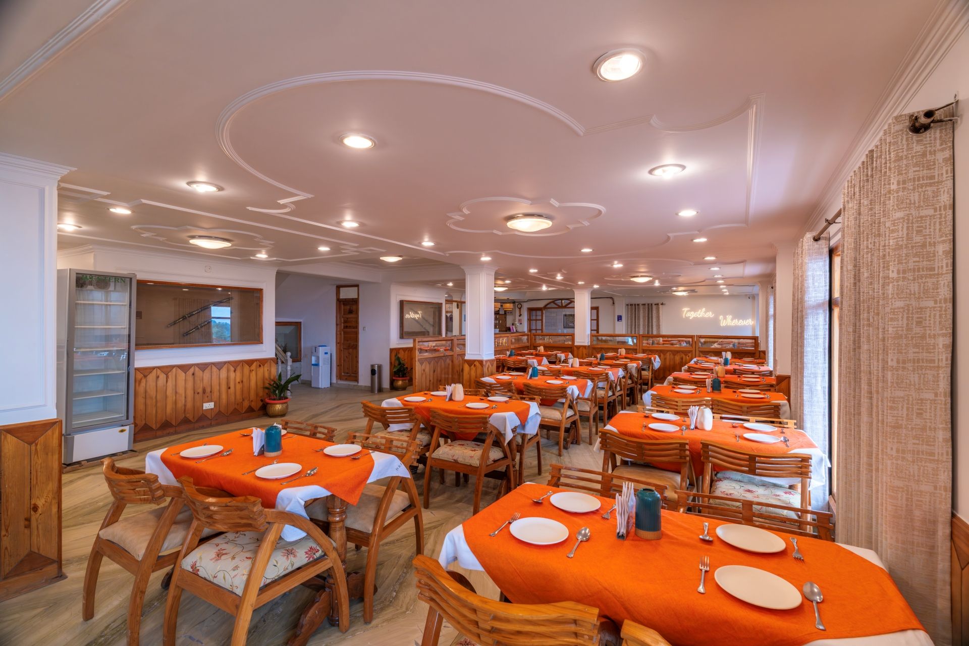 Un comedor amplio con varias mesas, cada una dispuesta con platos, cubiertos y manteles anaranjados.
