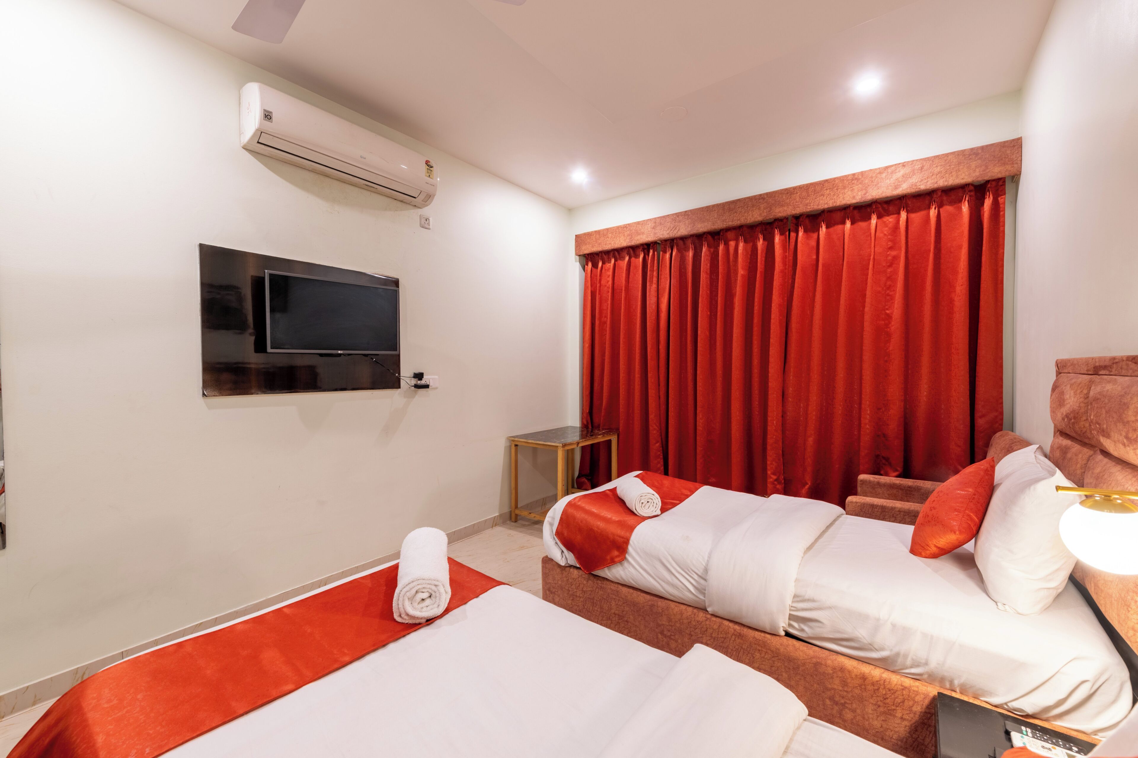 A hotel room with two beds, a TV, an air conditioner, and a window with red curtains.