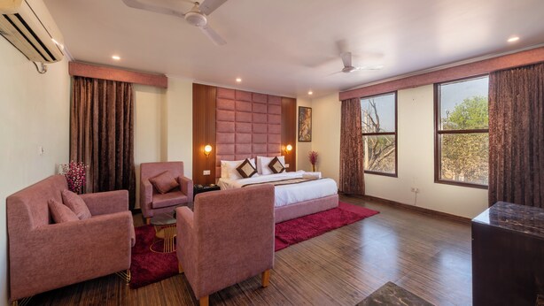 A hotel room with a bed, two chairs, a small table, a ceiling fan, and large windows. - La Comfort By shrpl (Gurugram)
