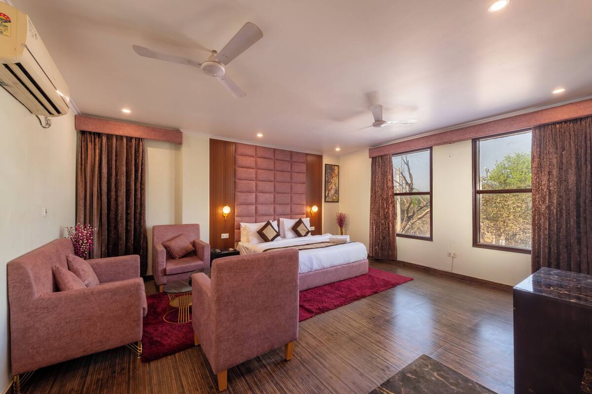 A hotel room with a bed, two chairs, a small table, a ceiling fan, and large windows.