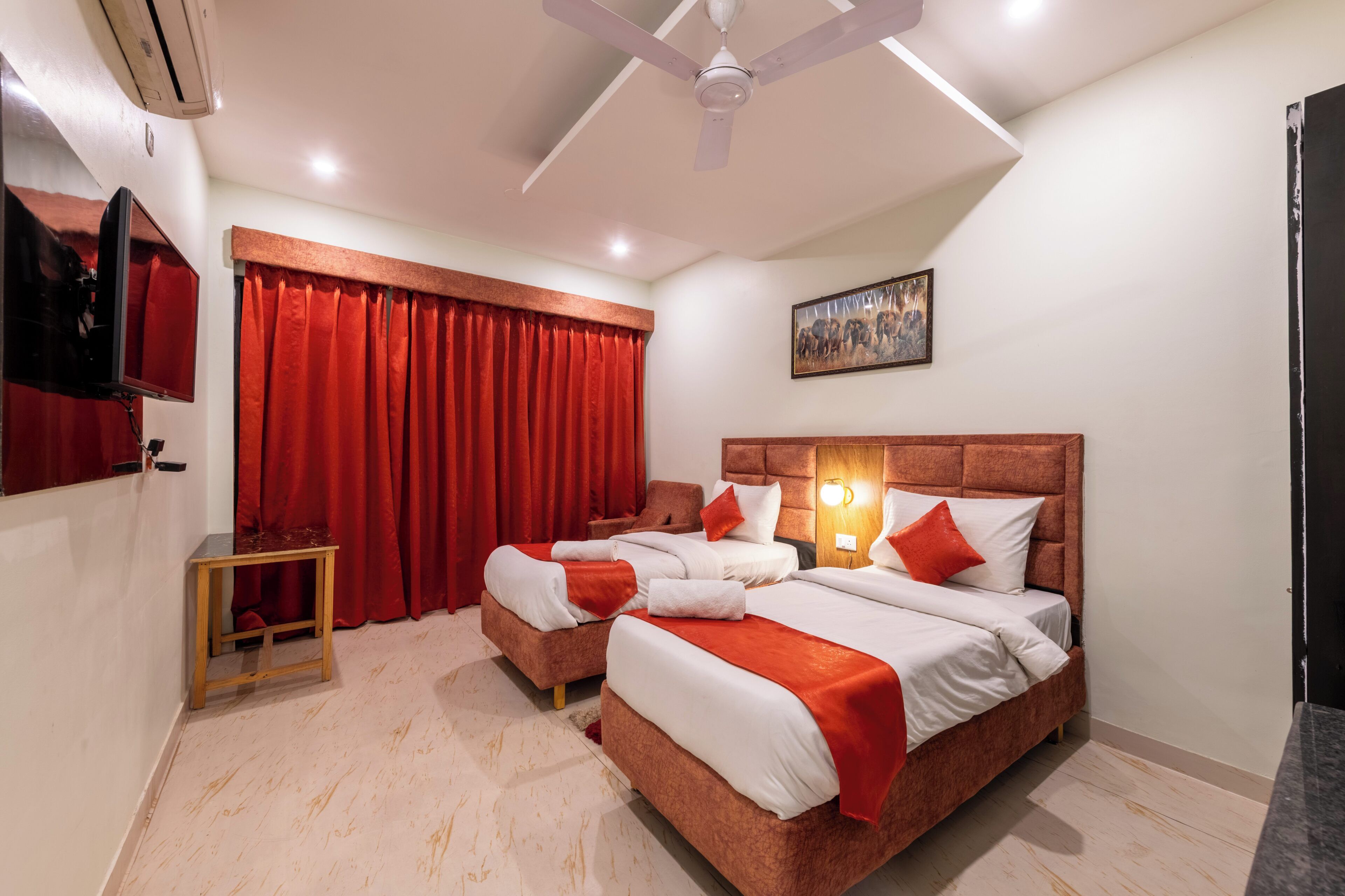 A hotel room with two beds, red curtains, a ceiling fan, a mounted TV, and a painting on the wall.