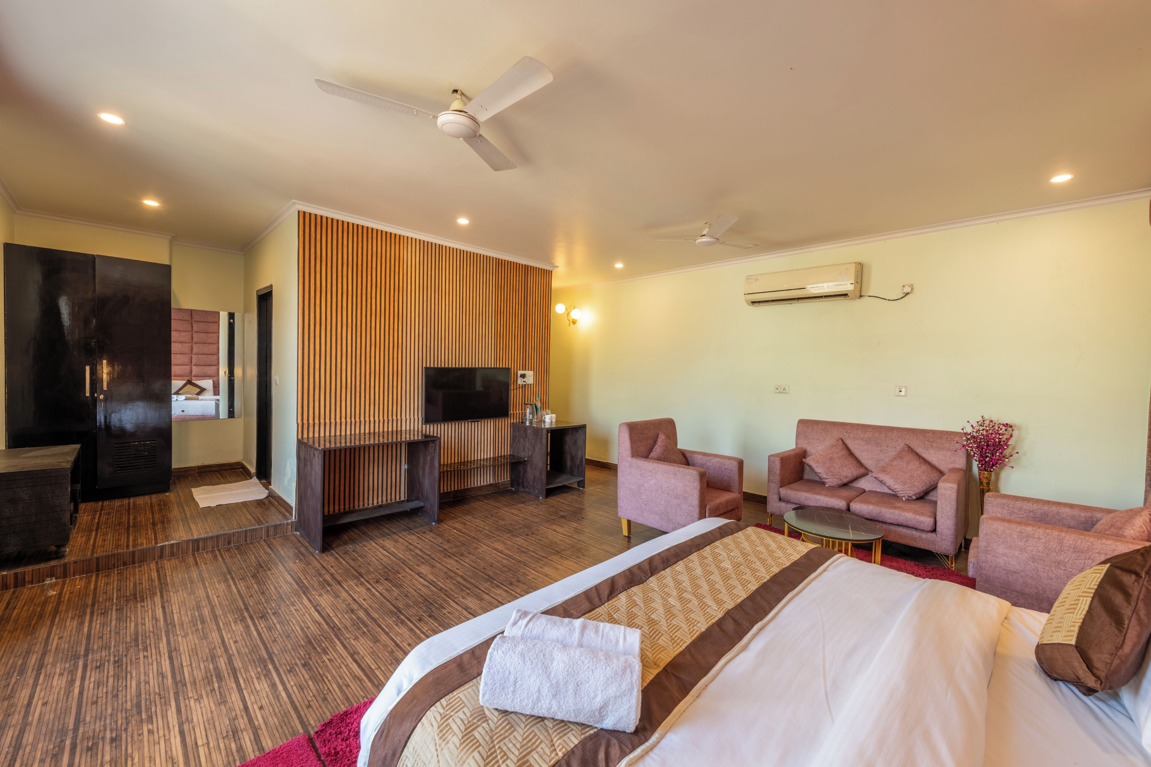 A hotel room with a bed, two sofas, a TV, a ceiling fan, and a wooden accent wall.