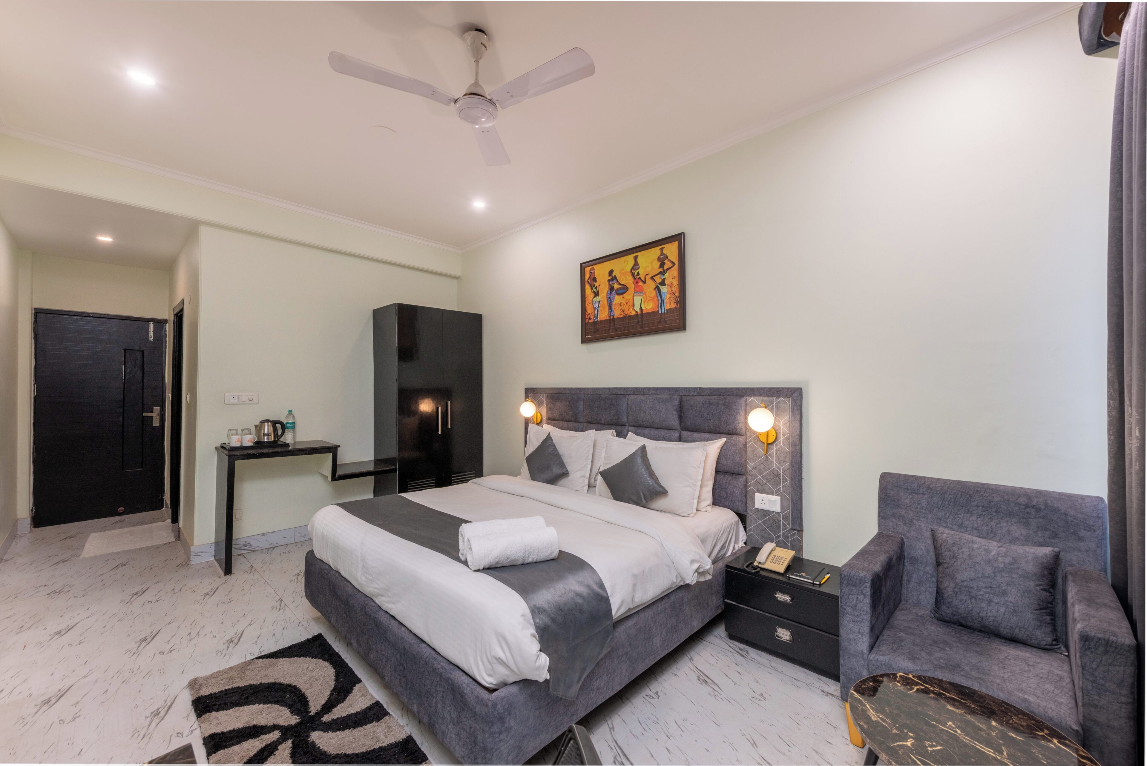 A modern hotel room with a bed, bedside table, chair, artwork, and a ceiling fan.