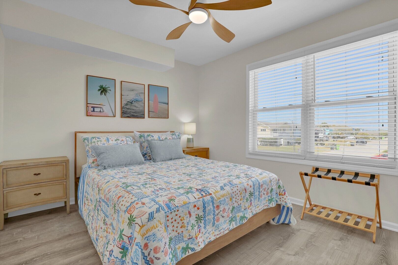 A bedroom with a bed, a nightstand, a bedside table, a ceiling fan, a window with blinds, and a view of the outside.