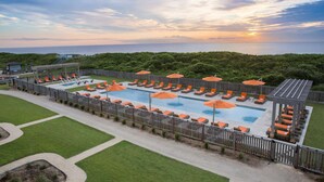 A pool area with multiple lanes, sun loungers, and umbrellas, surrounded by a wooden fence and greenery. - OBX Beach House + Pools. Sunsets & Ocean Path (Duck)