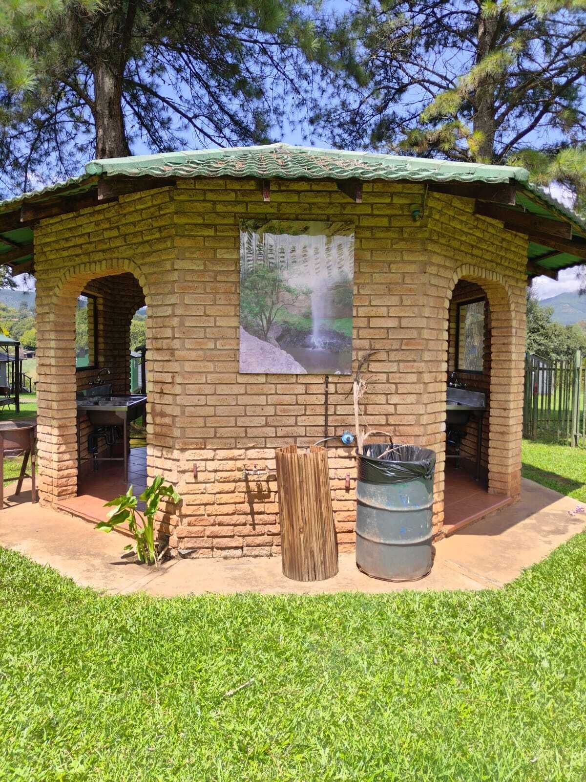 Un pabellón de ladrillo con techo verde, un mural de una cascada, una zona de barbacoa y un cubo de basura.