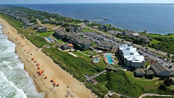An aerial view of a coastal resort with a beach, swimming pools, and multiple buildings.