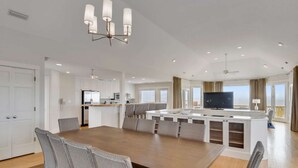 Un vaste espace de salle à manger avec une longue table et des chaises, une cuisine dotée de meubles blancs et un grand îlot central avec des sièges.