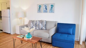 Una sala de estar con un sofá, un sillón azul, una mesa de centro de madera, una lámpara y tres cuadros colgados en la pared.