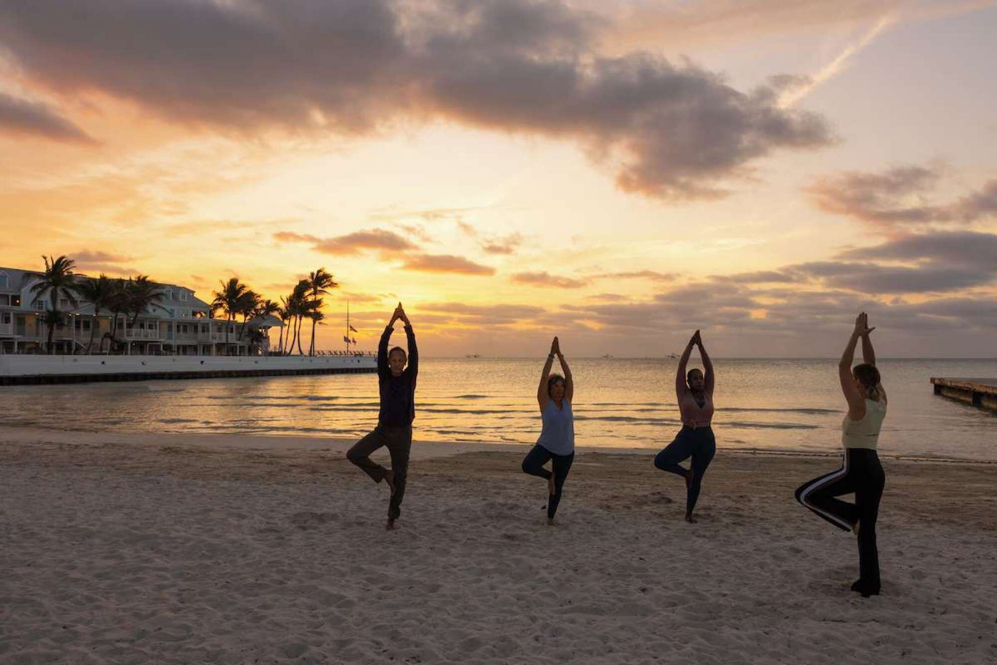 A beach with people practicing yoga, a sunset, and palm trees.