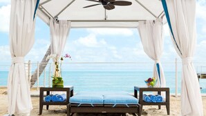 Un cabanon de plage doté d’un ventilateur de plafond, de deux petites tables en osier avec des serviettes, d’un banc avec des coussins et offrant une vue sur l’océan.