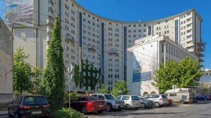 Ein mehrstöckiges Gebäude mit einer geschwungenen Fassade, parkenden Autos und Grünflächen.