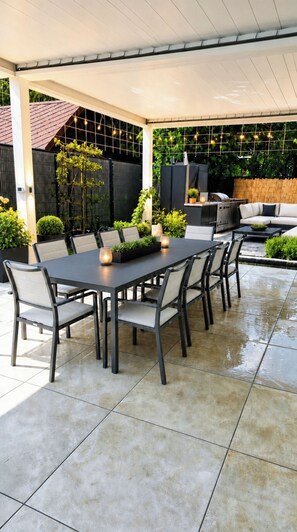 Eine Terrasse im Freien mit einem rechteckigen Tisch und Stühlen, umgeben von Blumentöpfen und einem Grill.