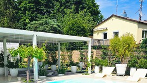 Ein Schwimmbecken mit überdachtem Bereich, umgeben von einem Garten mit verschiedenen Pflanzen und einer Rasenfläche.