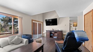 Ein Wohnzimmer mit Sofa, Esstisch, Wandfernseher und Blick ins Freie.