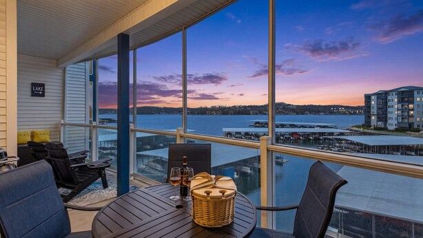 A balcony with a view of a lake and dock, furnished with chairs and a table, and a sign indicating 'LAKE'. - 2B 2B | Lake View | Seasonal Pool | Hot Tub |Grill (Lake Ozark)