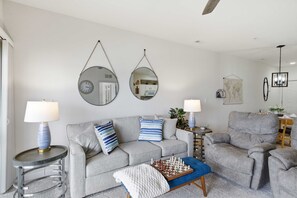 A living room with a grey sofa, two armchairs, a coffee table with chess pieces, a lamp, and two wall mirrors.