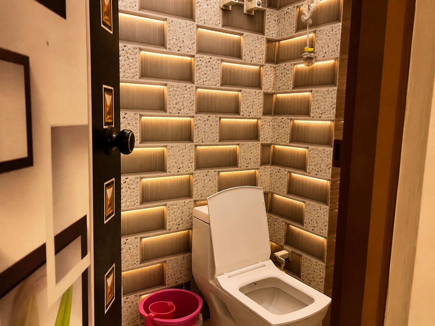 A modern bathroom with a unique tiled wall design, a toilet, and a red bucket.