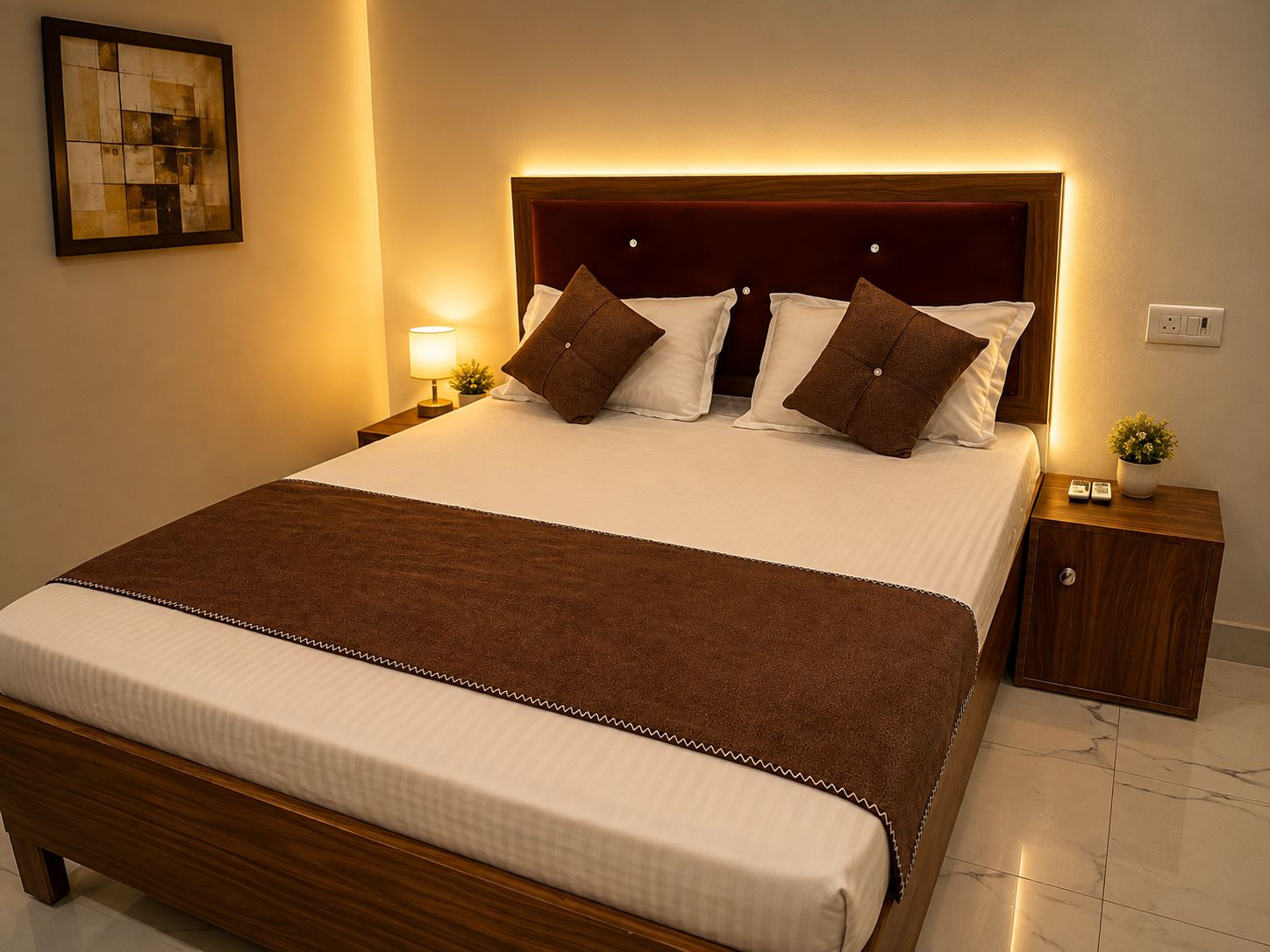 A neatly made bed with a brown headboard and white bedding, a bedside table with a lamp, and a small potted plant.