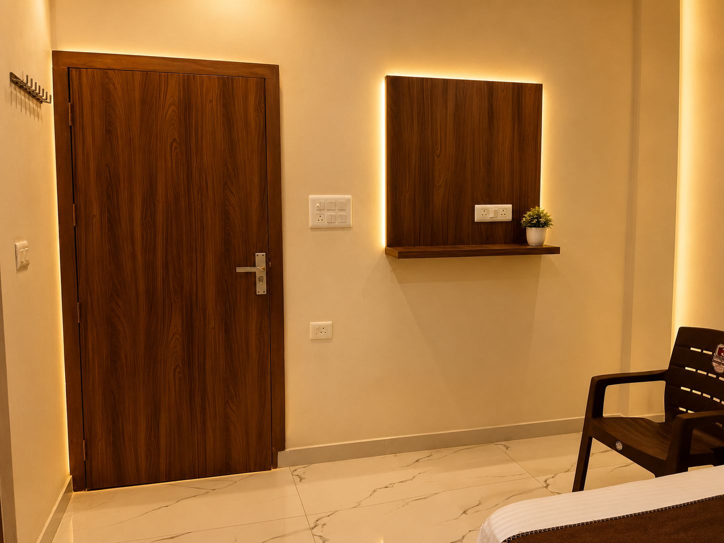 Una habitación con puerta de madera, repisa con una planta, una silla y cama con cabecera blanca.