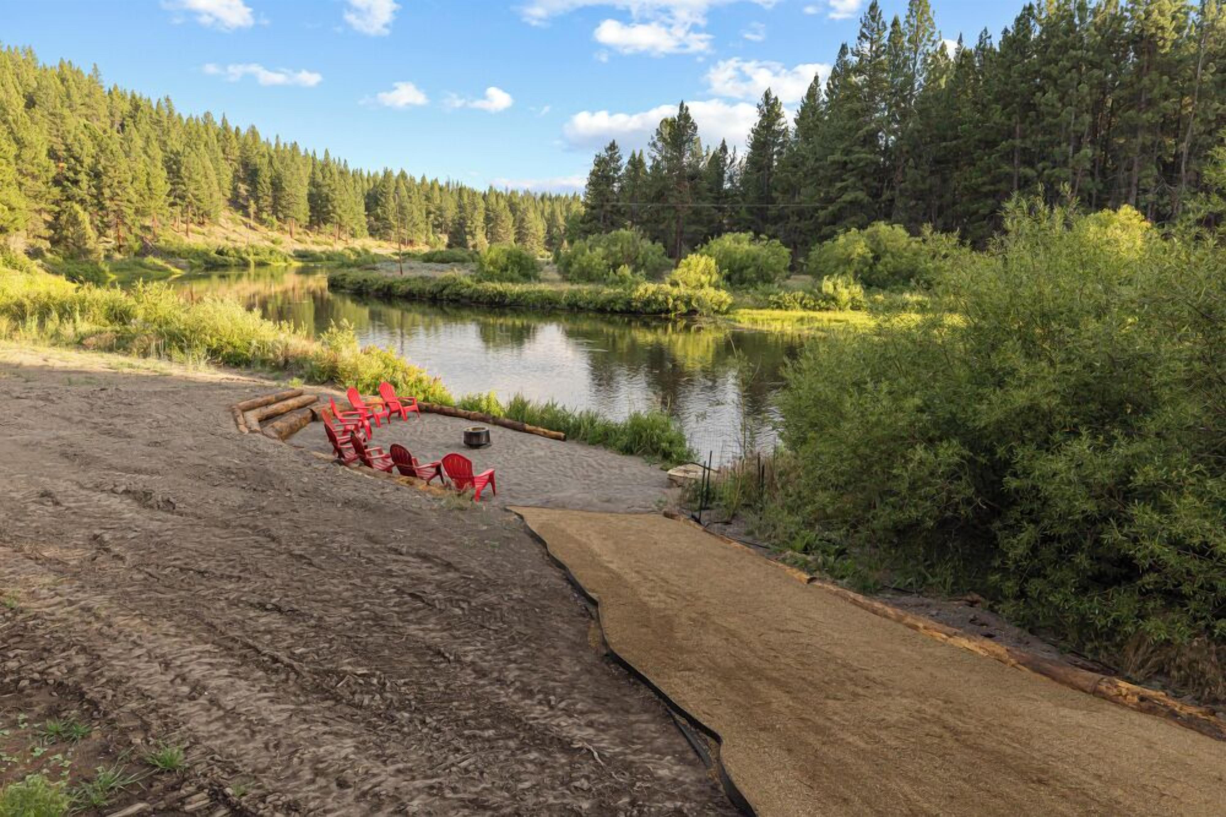A natural landscape with a lake, forest, and a sandy area with red chairs and a fire pit.