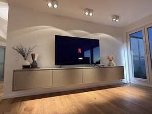 Ein modernes Wohnzimmer mit einem an der Wand montierten Flachbildfernseher, Holzboden und einem großen Fenster.