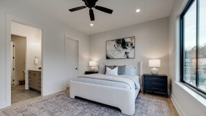 Een slaapkamer met een bed, twee nachtkastjes, een plafondventilator, een groot raam en een deur die naar een andere kamer leidt.