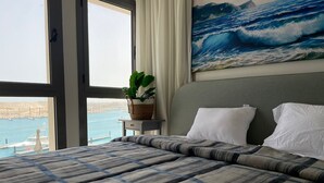 A hotel room with a bed, a view of the sea, a painting of waves, and a plant on a bedside table. - Penthouse 4 Bedrooms Marina2-14LR-305 (El Alamein)