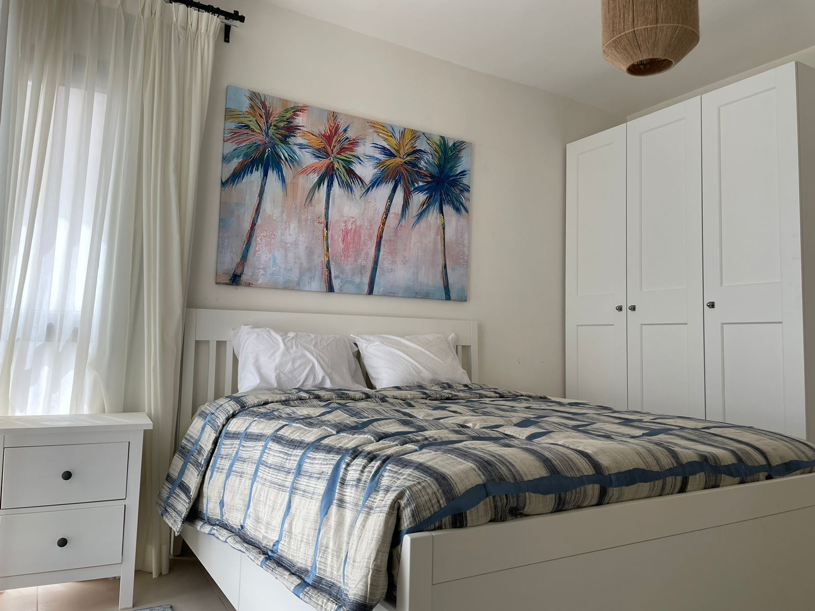 A bedroom with a bed, a nightstand, a wardrobe, and a painting of palm trees.