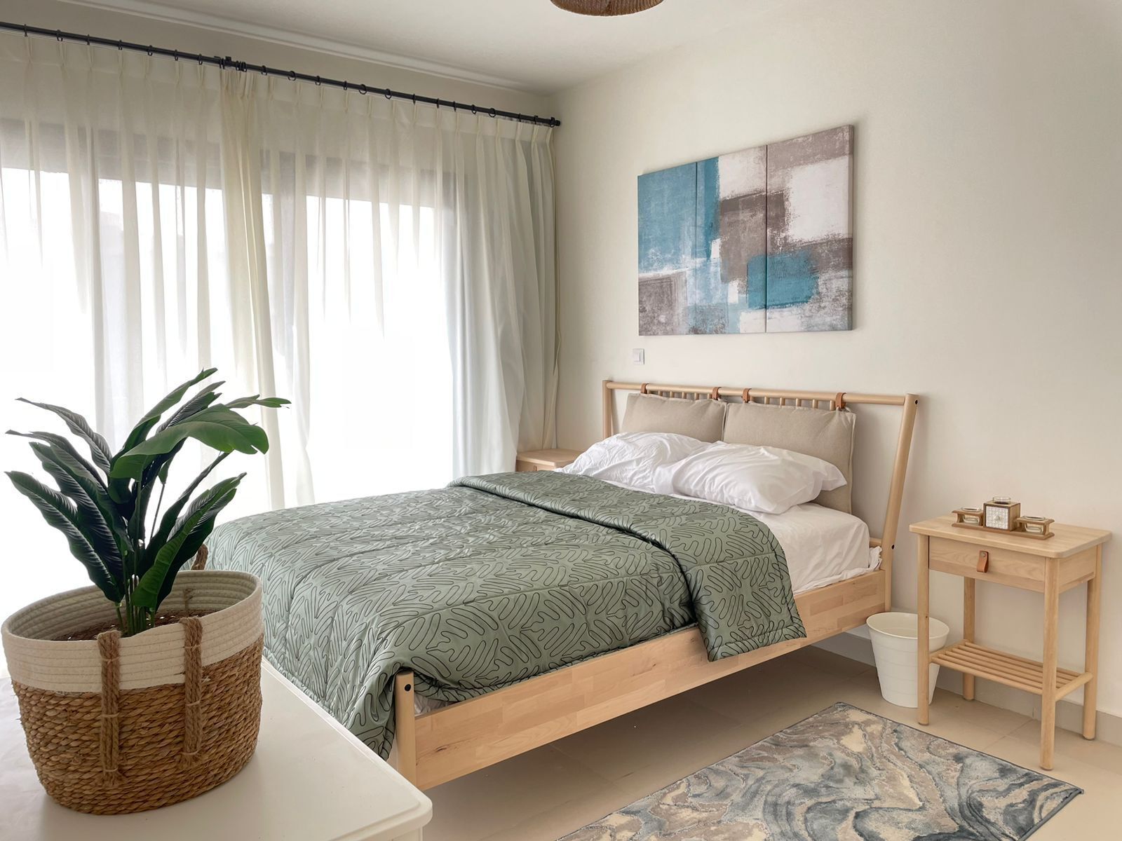 Un lit simple avec un cadre en bois, une table de chevet, une plante en pot, une œuvre d’art accrochée au mur et une fenêtre avec des rideaux légers.