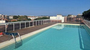 Una piscina en la azotea con terraza de madera, barandas metálicas y una vista despejada del paisaje urbano de fondo.