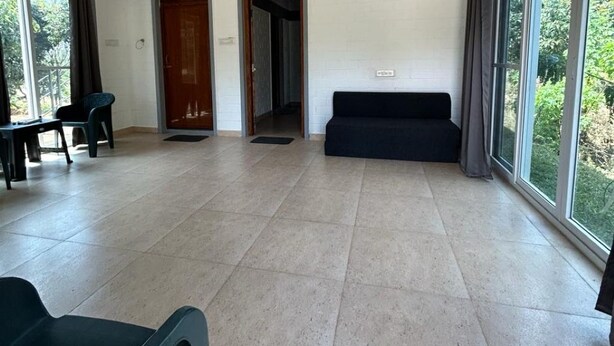 A spacious room with a ceiling fan, a black sofa, a dining table with chairs, and large windows offering a view of greenery. - Alphonso (Shoolagiri)