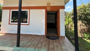 A covered outdoor area with a wooden floor, a white brick wall, and a door leading to an interior space. - Alphonso (Shoolagiri)
