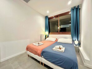 Ein Doppelbett mit zwei Kissen, ein Nachttisch mit Lampe, ein Fenster mit Vorhängen und eine Heizung.