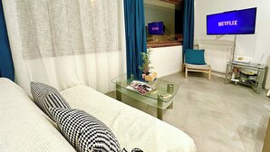 Un salon avec un canapé, une table basse en verre sur laquelle sont posés des magazines, un téléviseur diffusant Netflix, un fauteuil et une fenêtre avec des rideaux.